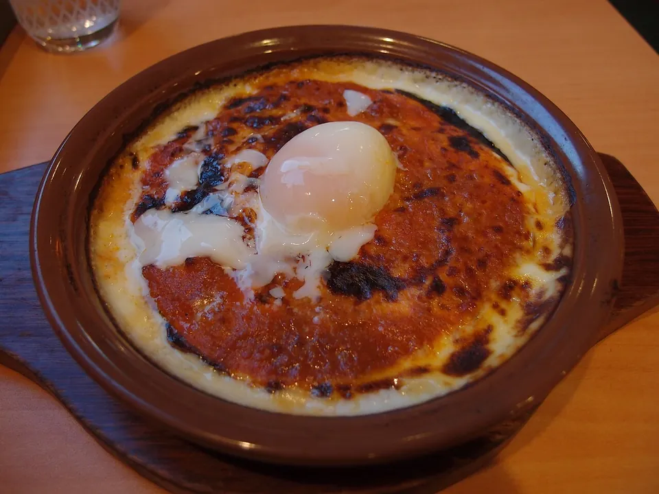 ドリアの実写写真（グラタン皿（陶製）で焼き上げた焼きドリア一皿＝焦げ目のついたチーズ/ソース面と皿全体がはっきり写り『ビーフシチューに見える』前回却下を回避。上の温泉卵はサイゼリヤ『ミラノ風ドリア＋卵』の現行トッピング変種（オリジナルは横浜ホテルニューグランド発祥のホワイトソース+ライス）。）
