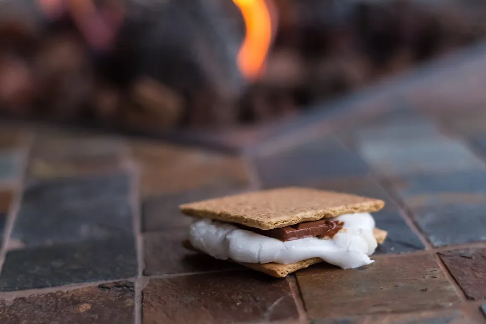 スモアの実写写真（米キャンプ文化の完成したスモア＝グラハムクラッカーで焼きマシュマロとチョコを挟んだ現行形が大写しの主役。焚き火を背景に手・人物は写り込まず。↺再挑戦=前回却下『人物や手の割合が大きい』を回避（スモア本体が大きく主題）。CC BY 2.0=帰属付き。）