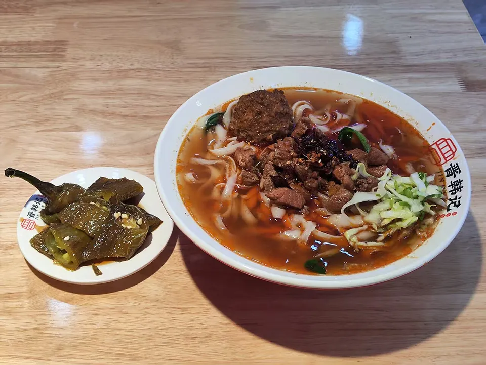 刀削麺の実写写真（刀削麺（山西の削り麺）の完成椀。太く不揃いに削られた麺が牛肉・辛味スープと共に盛られた現行の食堂供膳形（丼の店ロゴに『晋』＝山西の晋韓况）。CC0。）