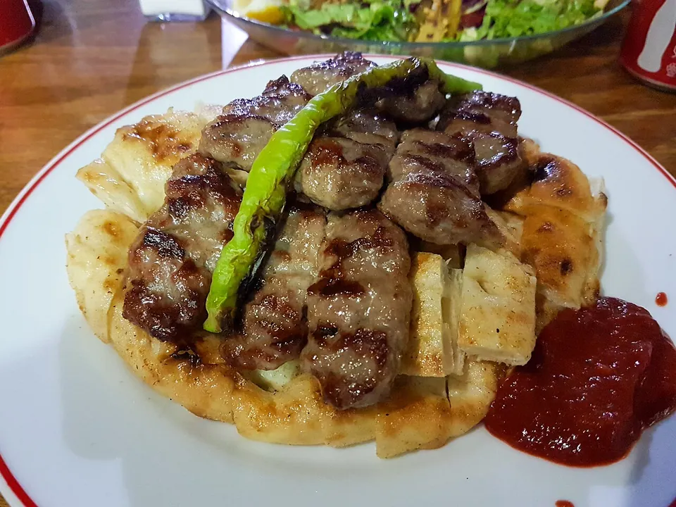 キョフテの実写写真（トルコ/アナトリアの köfte（焼き挽肉団子）の完成皿。トルコ・アクヒサル現地撮影のアクヒサル・キョフテ＝焼きキョフテの代表形（フラットブレッド・焼き青唐辛子・サルチャ添え）。総称キョフテの一変種として整合。）
