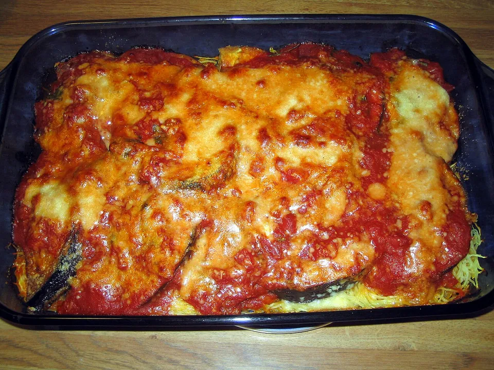 メランザーネ・アッラ・パルミジャーナの実写写真（現行形の完成皿（焼き上がり）。トマトソースで煮えたナスのスライスとチーズが全面に見え、ナスのパルミジャーナと判別できる。前回却下『写真がわかりにくい』（白いモッツァレラの塊が支配的で正体不明の接写）を解消＝赤いトマトソース主体で料理の正体が明瞭。撮影地=米国）