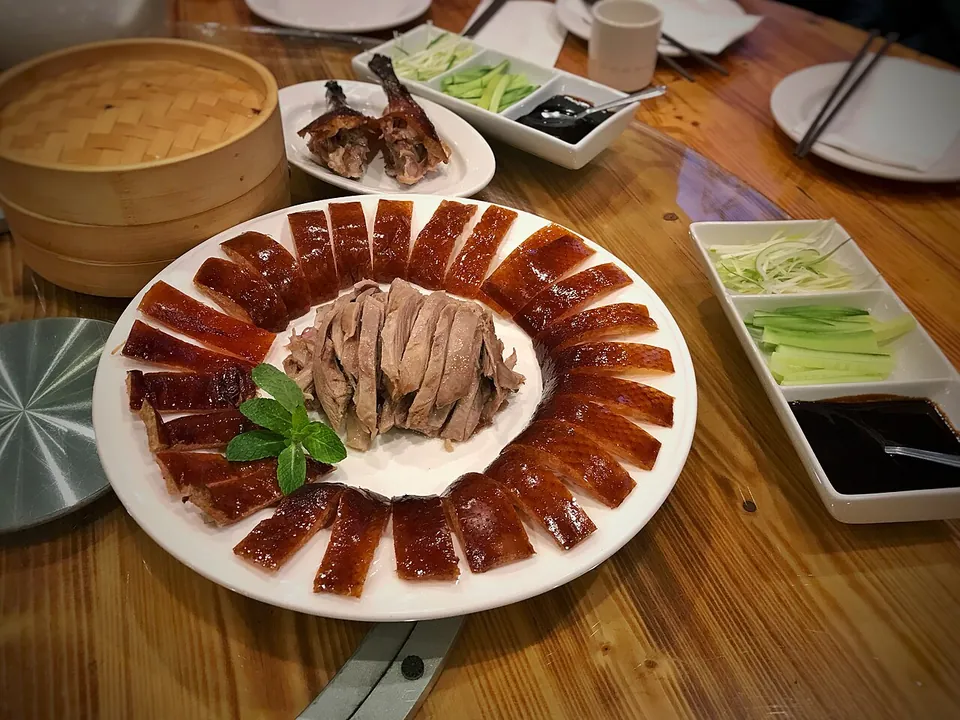 北京ダックの実写写真（スライスした皮・肉が皿の主役として大写しの完成供膳皿（薄餅の蒸籠・胡瓜・葱・タレ付き）。前回却下『主役が小さい』を回避＝ダック本体が中央で大きく写る一枚。撮影地は米国カリフォルニアの北京ダック専門店だが供膳形式は標準的な現行型。）
